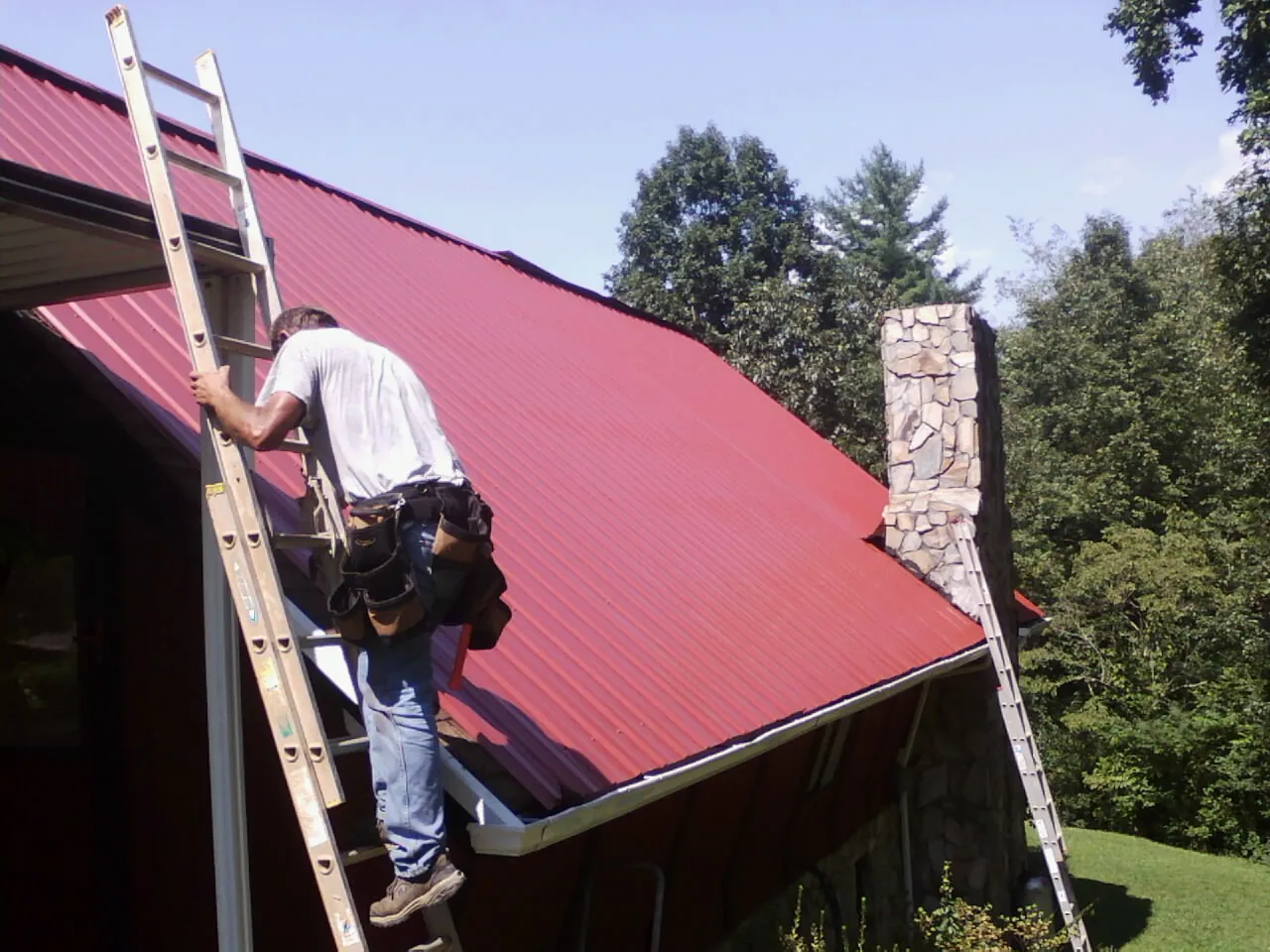Roofer on a ladder beside a red metal roof and stone chimney — Soffit & Fascia Repair by Roofing Brothers in Dawsonville